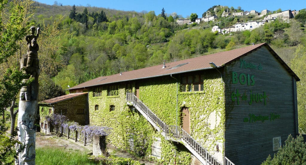 Maison du Bois et du Jouet en Montagne Noire La Maison du Bois et du Jouet en Montagne Noire, bâtiment couvert de lierre, avec sculpture totem et village en arrière-plan.